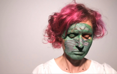 EL Putnam has pink hair and is wearing white. She is looking downwards with green paint on her face and overlay of child. 