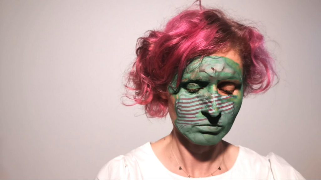 EL Putnam has pink hair and is wearing white. She is looking downwards with green paint on her face and overlay of child.